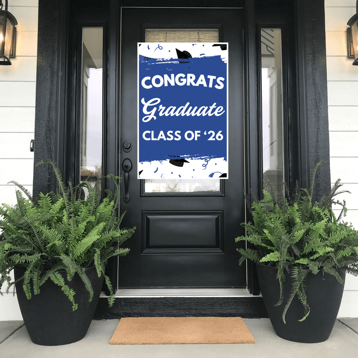 Blue and white graduation door banner with "Congrats Graduate Class of '26" text and decorative cap design, displayed on a black front door flanked by fern planters.