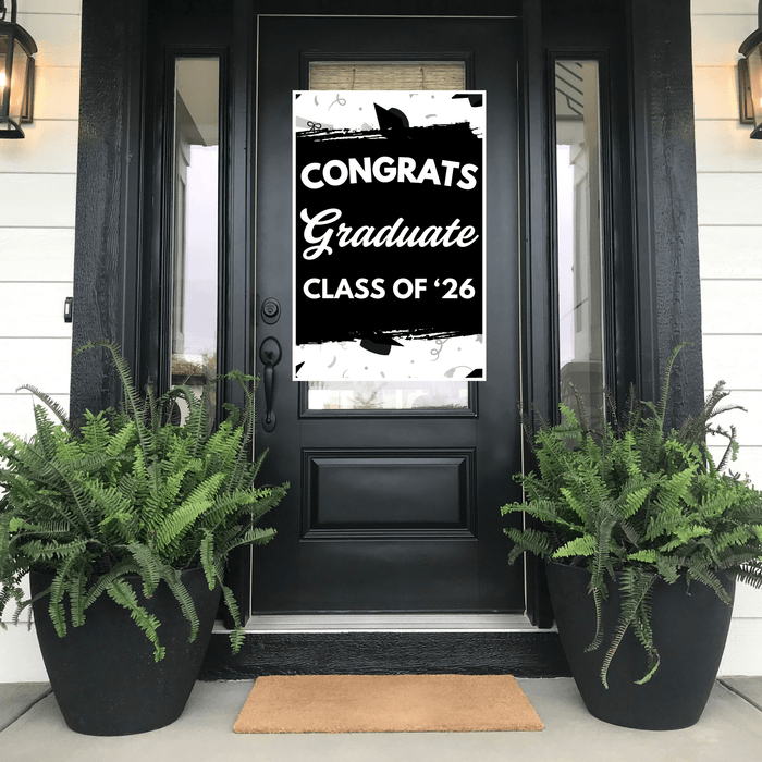 Black and white "Congrats Graduate Class of '26" door banner displayed on a black front door with ferns in black planters on either side.