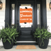 Orange and white graduation door banner displaying "CONGRATS Graduate CLASS OF '26" with mortarboard graphics, hung on a black front door flanked by fern planters.