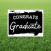 Black and white lawn yard sign with "CONGRATS" in bold capital letters and "Graduate" in white script font on a black brushstroke background, displayed on green grass.