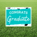 Turquoise and white yard sign with "CONGRATS Graduate" text in white lettering, decorated with black graduation cap illustrations and paint splatter accents.