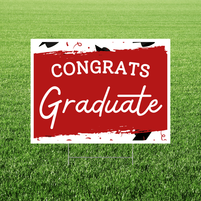 Red and white yard sign with black graduation cap illustrations and white text reading "CONGRATS Graduate" displayed on green grass.