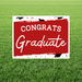 Red and white yard sign with black graduation cap illustrations and white text reading "CONGRATS Graduate" displayed on green grass.