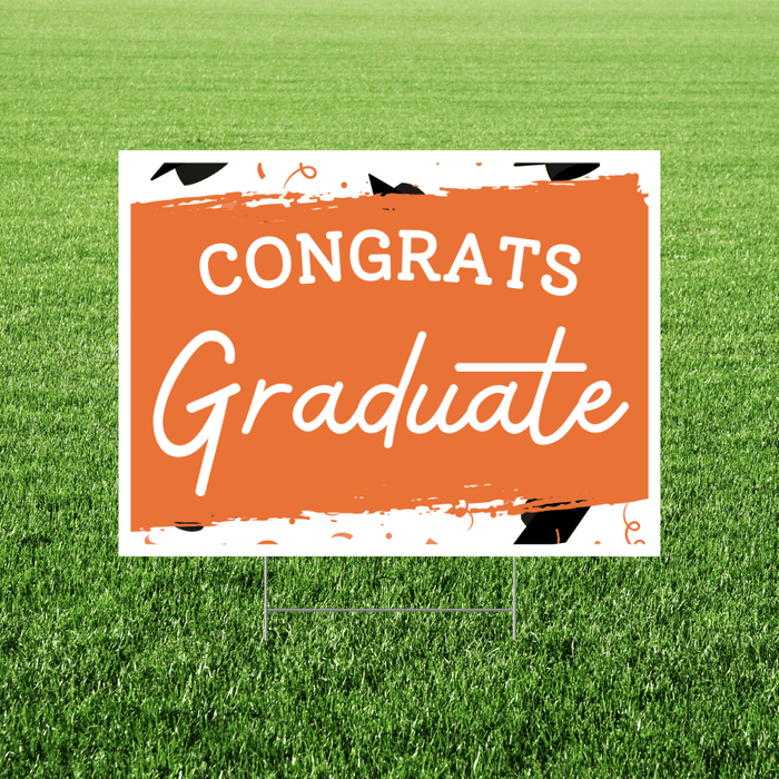 Orange and white yard sign with "CONGRATS Graduate" text and decorative graduation cap elements, displayed on grass.