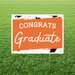 Orange and white yard sign with "CONGRATS Graduate" text and decorative graduation cap elements, displayed on grass.
