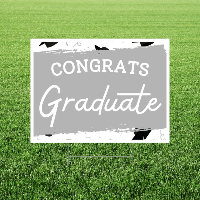 White yard sign with gray center panel featuring "CONGRATS" in capital letters and "Graduate" in script font, decorated with black graduation cap illustrations, displayed in grass.