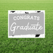 White yard sign with gray center panel featuring "CONGRATS" in capital letters and "Graduate" in script font, decorated with black graduation cap illustrations, displayed in grass.