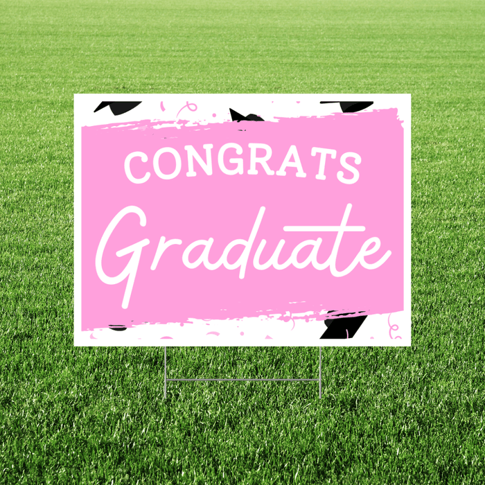 Pink yard sign with white text reading "CONGRATS Graduate" and black graduation cap decorations, displayed in grass.