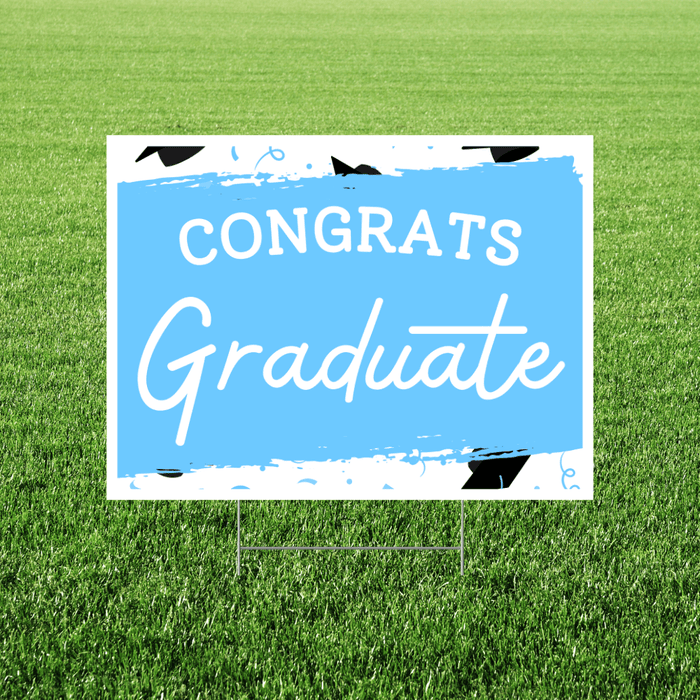 Light blue and white yard sign with black graduation cap illustrations and white text reading "CONGRATS Graduate" displayed on green grass.