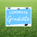 Light blue and white yard sign with black graduation cap illustrations and white text reading "CONGRATS Graduate" displayed on green grass.