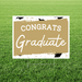Outdoor yard sign with gold and white color scheme displaying "CONGRATS Graduate" text with graduation cap icons, mounted on a metal stake in grass.