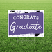 Purple and white yard sign with "CONGRATS Graduate" text and graduation cap motifs, mounted on a metal stake in grass.