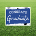 Blue and white yard sign with "CONGRATS Graduate" text and decorative black paint splatter accents, displayed on green grass.
