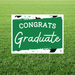 Green and white yard sign with "CONGRATS Graduate" text and graduation cap design, mounted on a stake in grass.