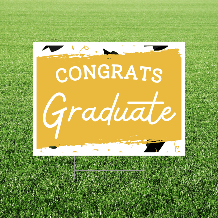 Yellow and white yard sign with "CONGRATS Graduate" text and graduation cap graphics, displayed on green grass.