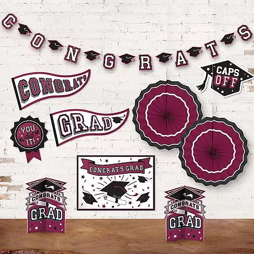 Maroon and black graduation decoration set includes a "Congrats" banner with mortarboard cutouts, two large paper fan decorations, two "Congrats Grad" boxes, and various themed cutouts and stickers on white brick background.