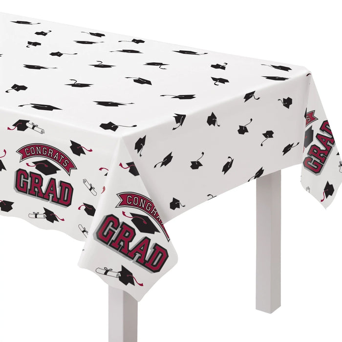 White plastic tablecloth with scattered black graduation caps and maroon ribbons, featuring "Congrats Grad" text in maroon and gray, draped over a white table.