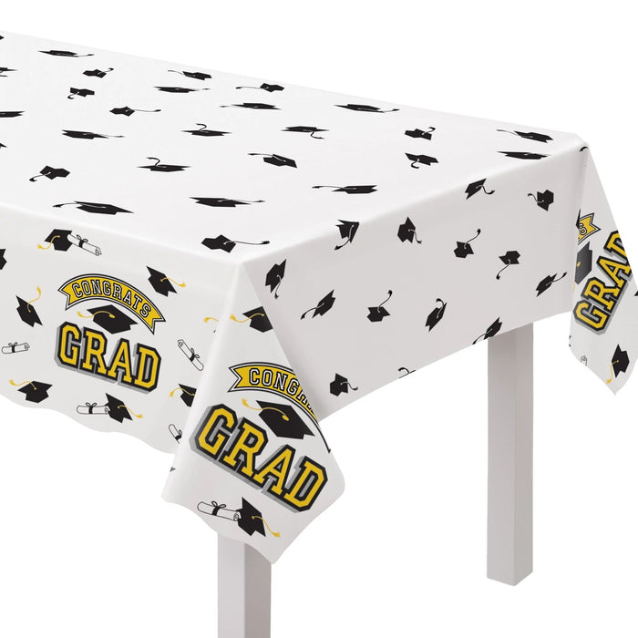 White plastic tablecloth with scattered black graduation caps and diplomas, yellow and black "Congrats Grad" banners, draped over a white table.