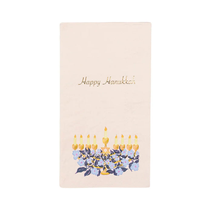 Cream-colored guest towel napkin featuring "Happy Hanukkah" text in gold script at the top and an illustrated menorah with nine golden candles and blue flowers at the bottom.
