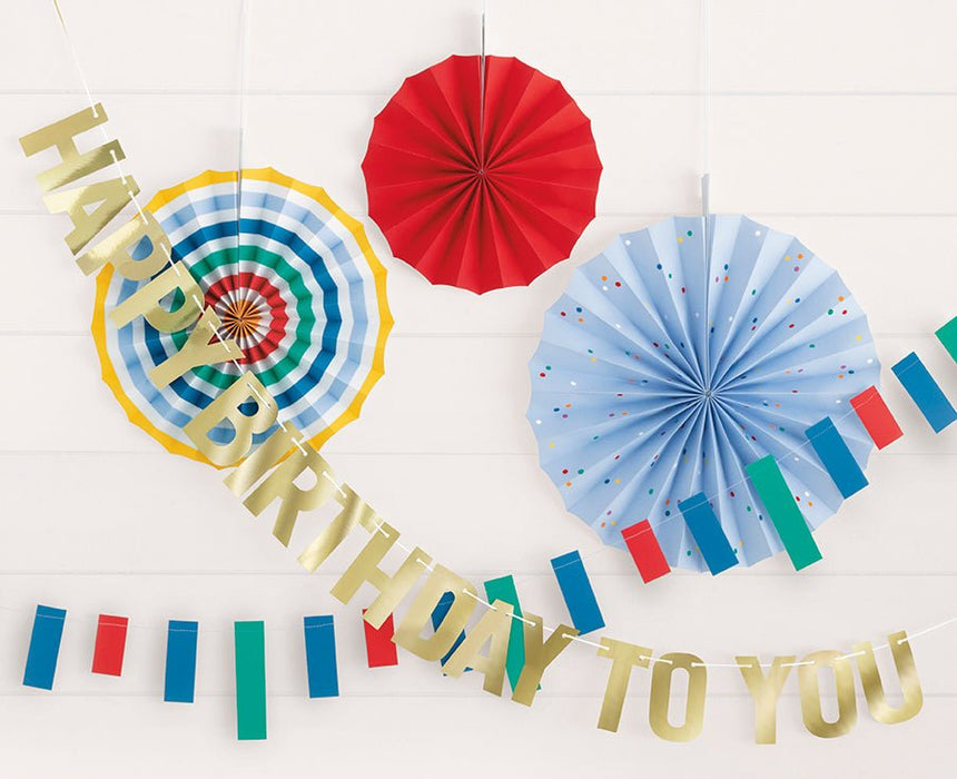 Gold "Happy Birthday to You" garland with colorful flag bunting, red accordion fan, light blue polka dot fan, and yellow striped circular fan displayed against white brick backdrop.