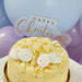 Clear acrylic cake topper with gold lettering reading "Happy Christening" displayed on a pale yellow frosted cake decorated with white chocolate discs and almond flakes against a purple balloon background.