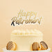 Gold and black "Happy Retirement" cake topper with cloud-shaped backing on a white frosted cake decorated with piped frosting and chocolate-drizzled cookies.