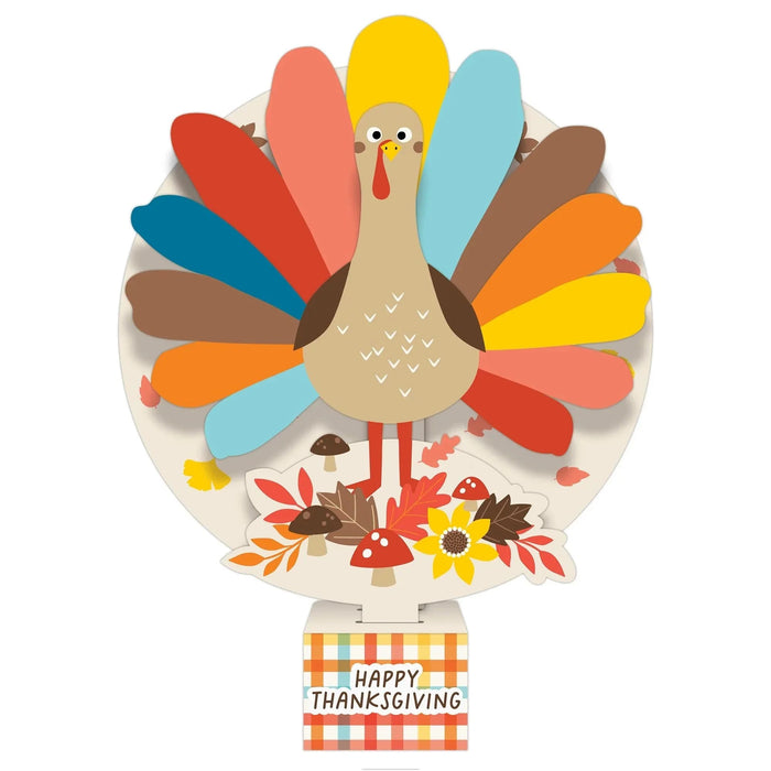 Colorful paper pop-up centerpiece featuring a turkey with multicolored feathers in red, blue, yellow, orange, cyan, and brown, surrounded by autumn leaves, mushrooms, and flowers on a cream base labeled "Happy Thanksgiving" in a gingham checkered banner.