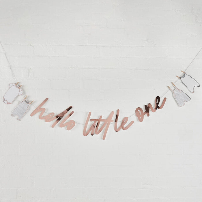 Rose gold script banner reading "hello little one" with small white onesie and dress decorations hanging from strings against a white brick wall.