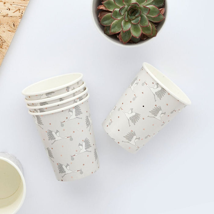 Four cream-colored paper cups with gray stork and coral dot patterns stacked and displayed on a white surface beside a succulent plant.