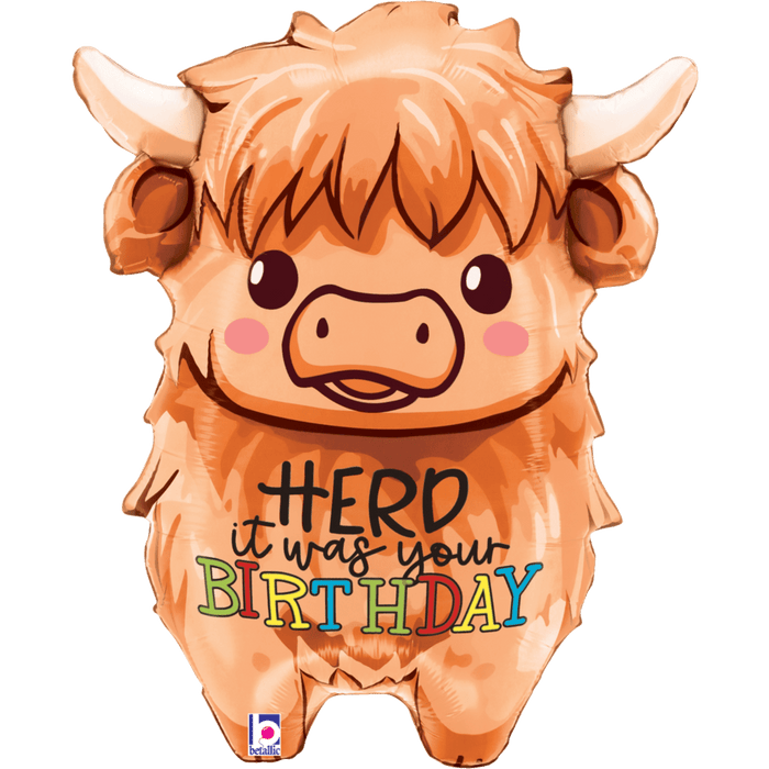 Orange mylar balloon shaped like a fluffy Highland cow with horns, brown eyes, pink cheeks, and text reading "it HERD it was your BIRTHDAY" in colorful letters across its body.