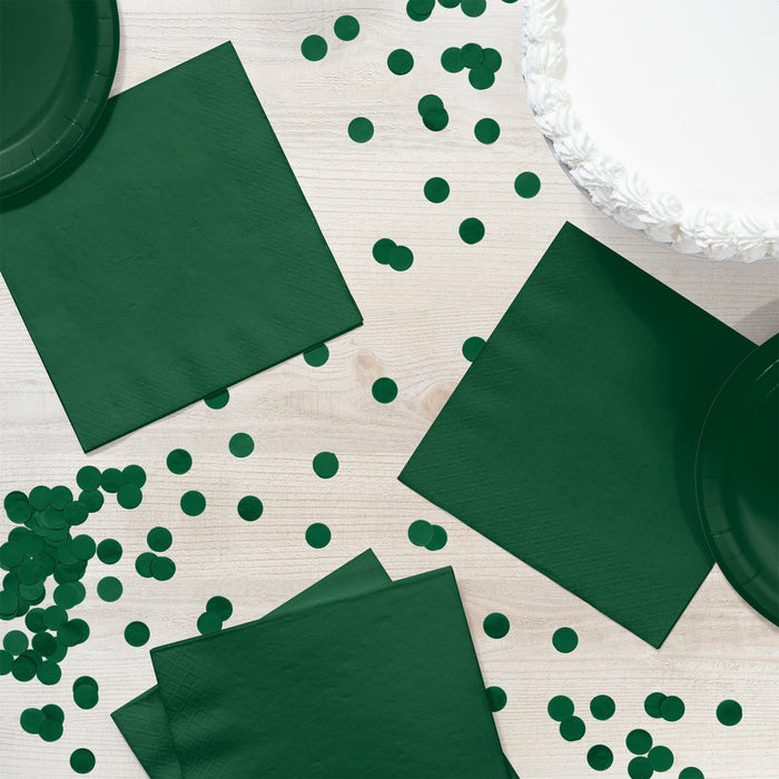 Hunter green paper napkins and coordinating party supplies arranged on a white wooden surface with scattered dark green circle confetti.