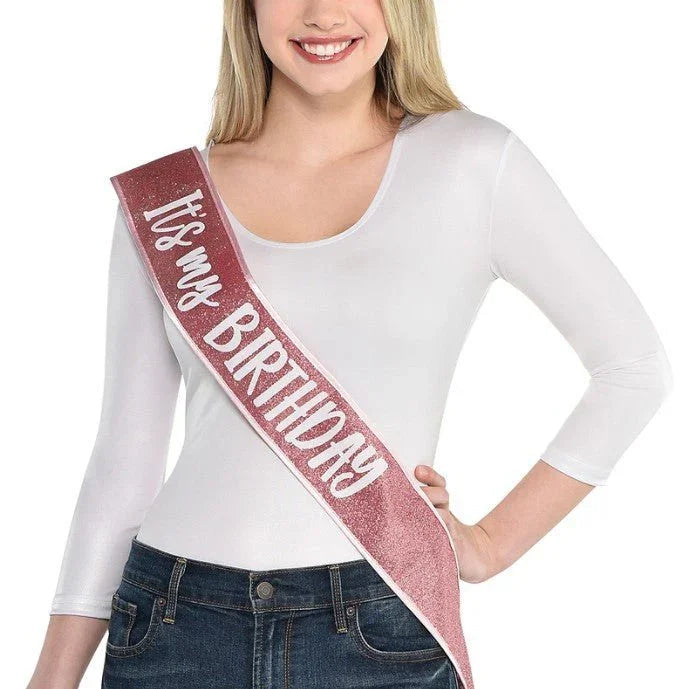 Smiling woman wearing a white long-sleeve top and jeans with a burgundy glitter sash diagonally across her body that reads "It's My Birthday" in white text.
