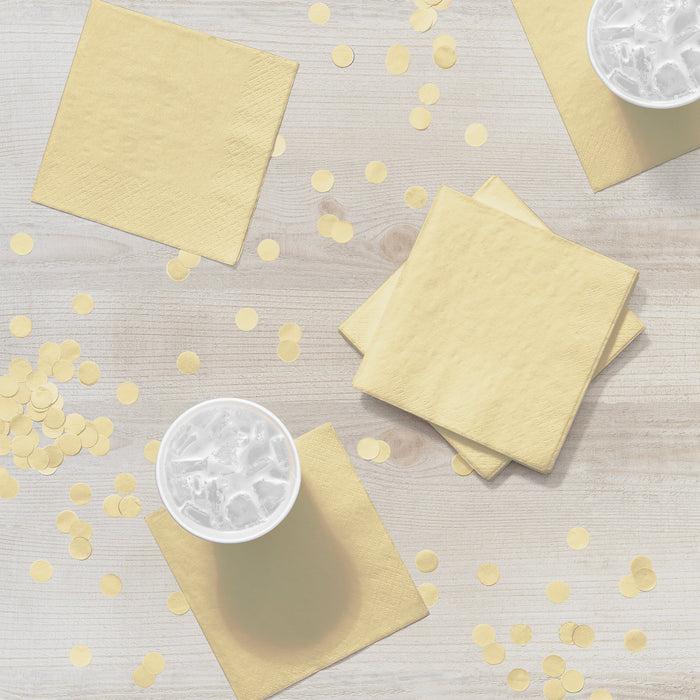 Ivory paper napkins arranged on a light wooden surface with small round confetti pieces scattered between them, featuring stacked napkins and two white cups with ice.