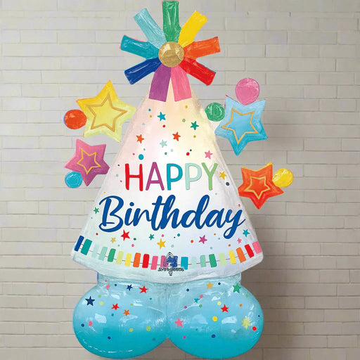 Colorful Happy Birthday party hat shaped foil balloon with rainbow ribbons on top, stars and dots design, standing 27 inches tall against a white brick wall.