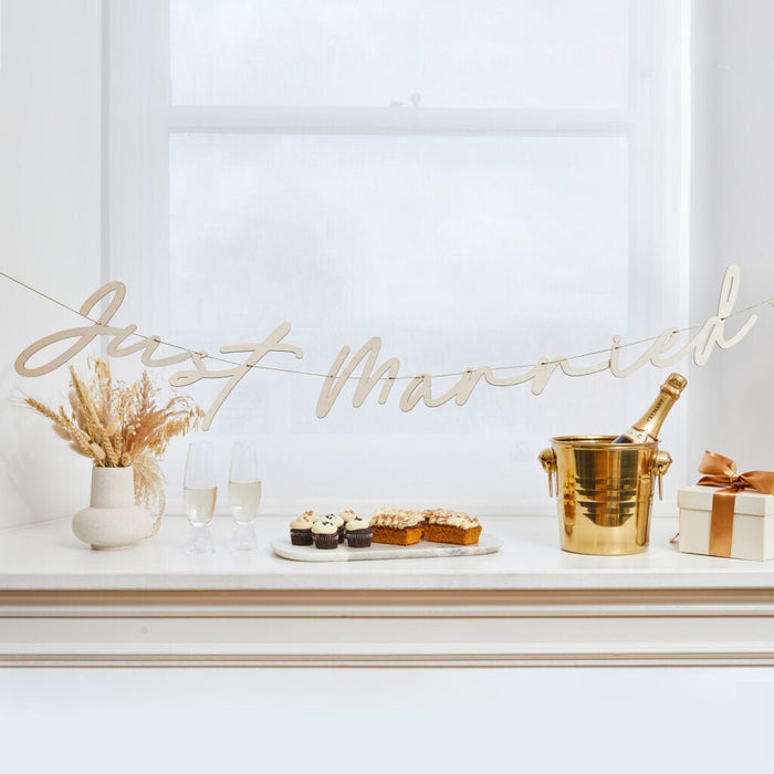 Wooden "Just Married" banner in natural beige script lettering hanging above an elegant white mantelpiece display with champagne, cupcakes, cake slices, pampas grass, and gold accents.