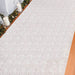 White lace patterned aisle runner displayed on wooden flooring with decorative greenery and white candles arranged along the top edge.