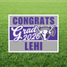 Purple and gray yard sign with white bull logo displaying "Congrats Grad 2026 LEHI" text on grass background.