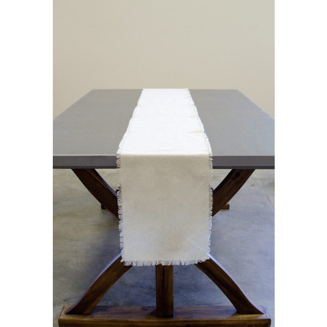 Cream linen table runner with frayed edges draped down the center of a dark wood dining table with X-shaped legs.