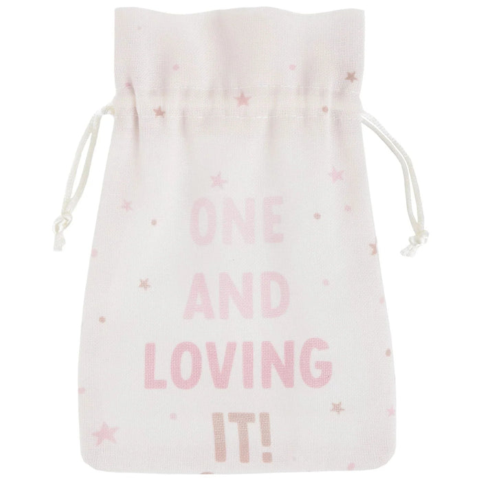 White drawstring pouch with pink stars and text reading "ONE AND LOVING IT" in pink letters scattered across the front.