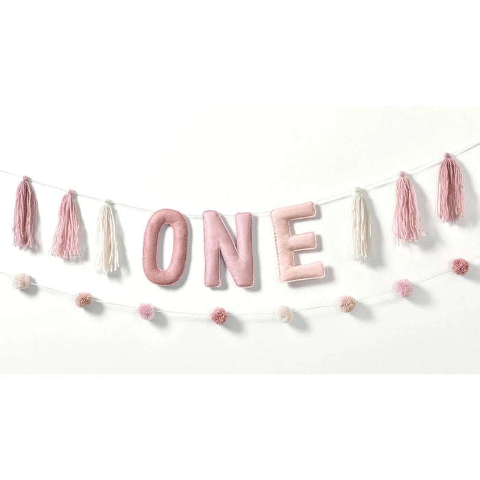 Plush banner kit featuring large padded letters spelling "ONE" in pink and cream, accented with pink and cream tassel garland and pom-pom garland strands for first birthday decoration.