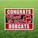 Red and black yard sign with white text reading "CONGRATS Grad 2026 BOBCATS" and a red paw print, displayed on green grass.