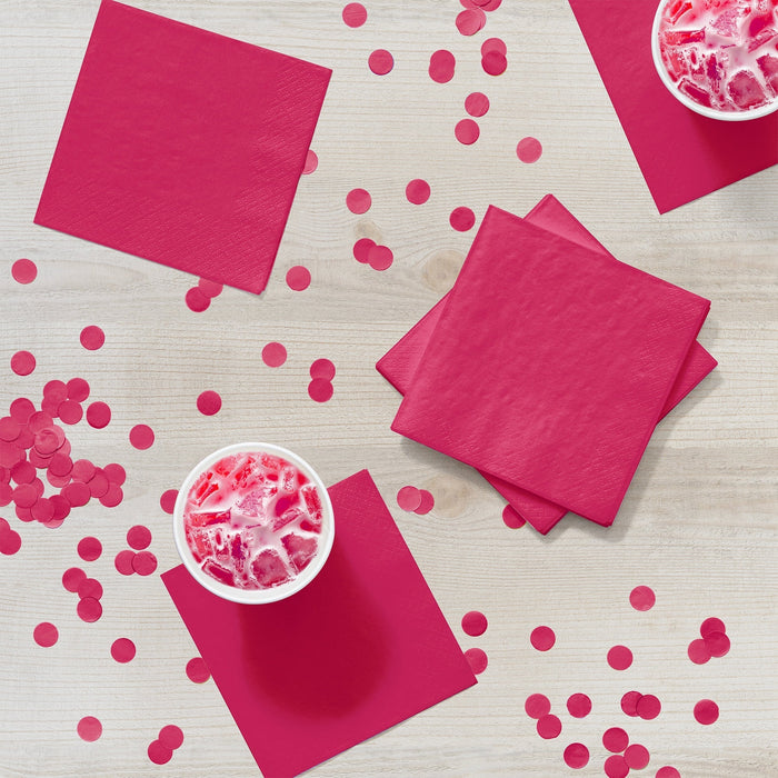 Bright magenta paper napkins arranged on a white wooden surface with pink confetti and two bowls of pink frozen dessert.