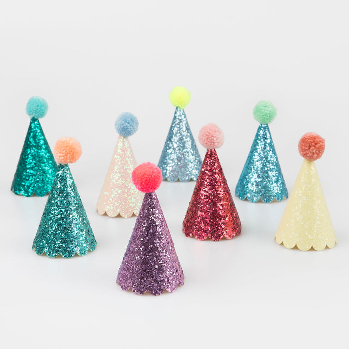 Assorted glitter mini cone party hats in multiple colors including pink, purple, teal, red, yellow, and blue, each topped with a pom-pom.