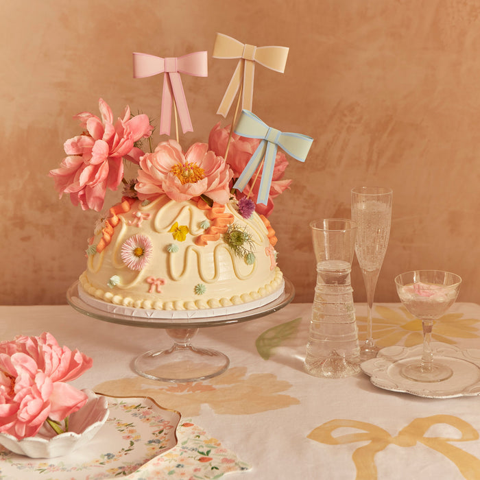 Pastel Bow Cake Toppers featuring peach, yellow, and mint paper bows on wooden picks decorating a dome cake with flowers.
