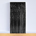 Black metallic fringe curtain with vertical strands hanging from a white header strip, displayed leaning against a white wall on a wooden floor.