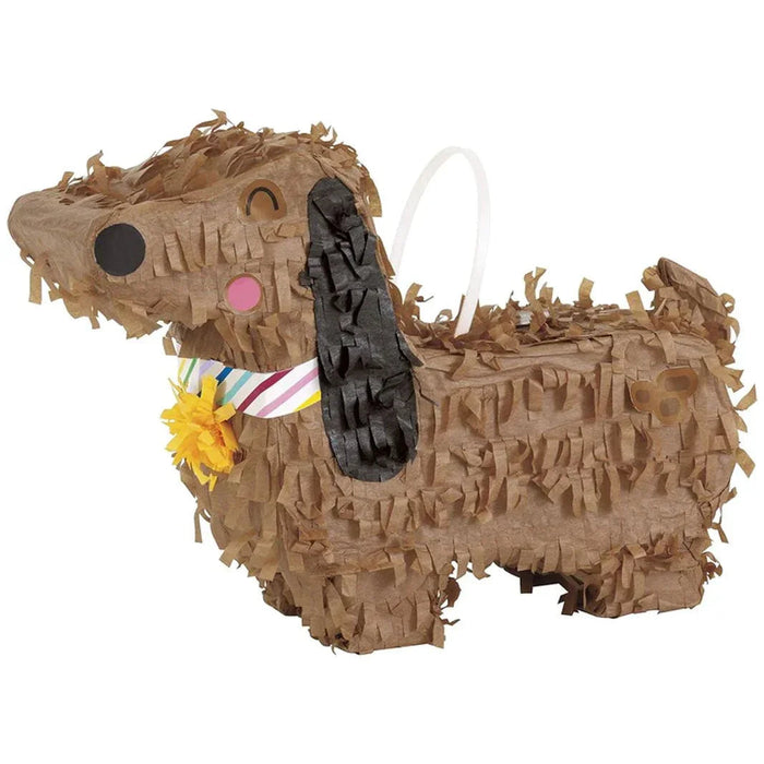 Tan cardboard piñata shaped like a dachshund with a black oval nose, pink circle cheek, black ear, colorful striped ribbon bow, yellow flower accent, and white hanging loop.