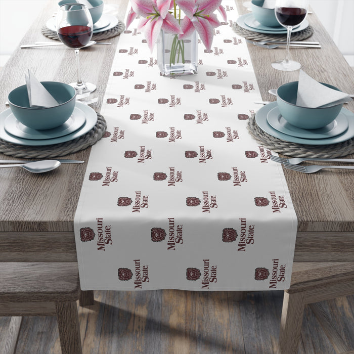 White table runner with repeating Missouri State logo pattern in burgundy, displayed on a wooden table set for two with teal bowls, wine glasses, and pink lilies in a vase.