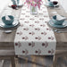 White table runner with repeating Missouri State logo pattern in burgundy, displayed on a wooden table set for two with teal bowls, wine glasses, and pink lilies in a vase.