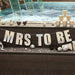 Iridescent white letter banner spelling "MRS TO BE" strung on twine above a dark wooden pier, decorated with heart-shaped sunglasses, silver disco balls, and metallic cups with an ocean backdrop.