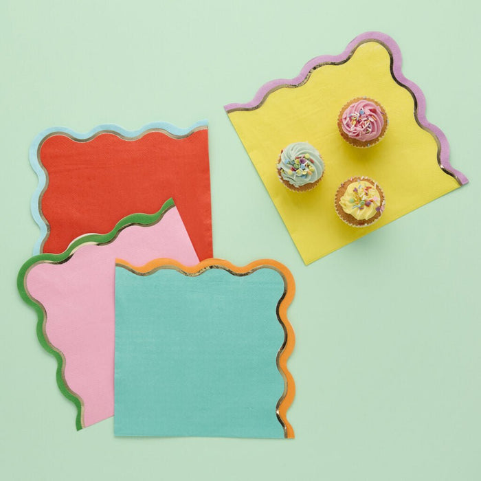 Colorful die-cut napkins with scalloped edges and metallic trim in red, pink, turquoise, and yellow, displayed with three decorated cupcakes on a pale green background.
